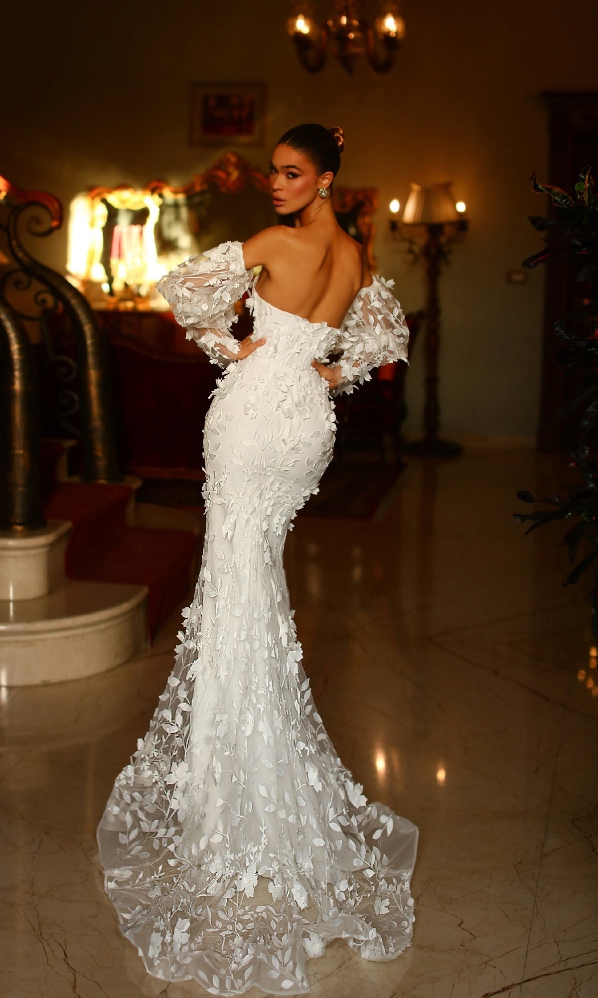 A woman in the Tina Holly TW121W puff-sleeve white formal gown with floral appliqués and a long train stands indoors, looking over her shoulder.